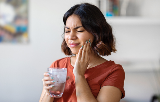 Woman holding glass of water touching cheek in pain, suggesting tooth sensitivity or dental discomfort.