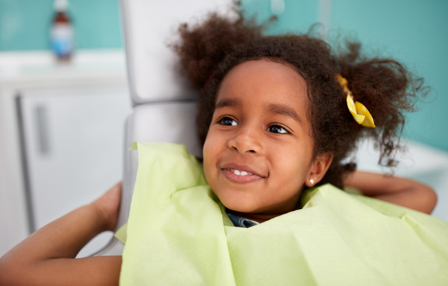 Young child in yellow shirt smiling at dental office with turquoise wall background.