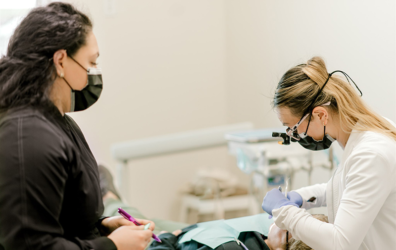 Dental professional in white coat examines patient's teeth in clinical treatment room.