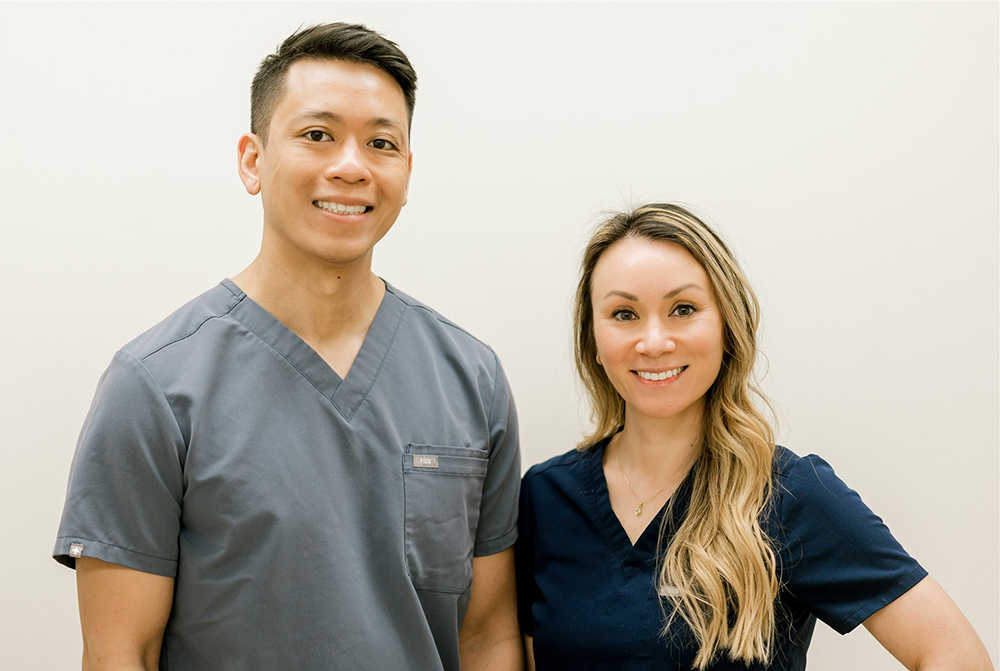 Two dental professionals smiling at camera in clinical office setting wearing scrubs.