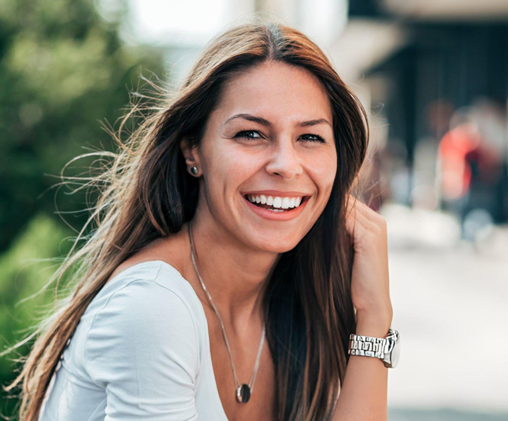 Smiling woman with long dark hair wearing light blue top outdoors in urban setting.