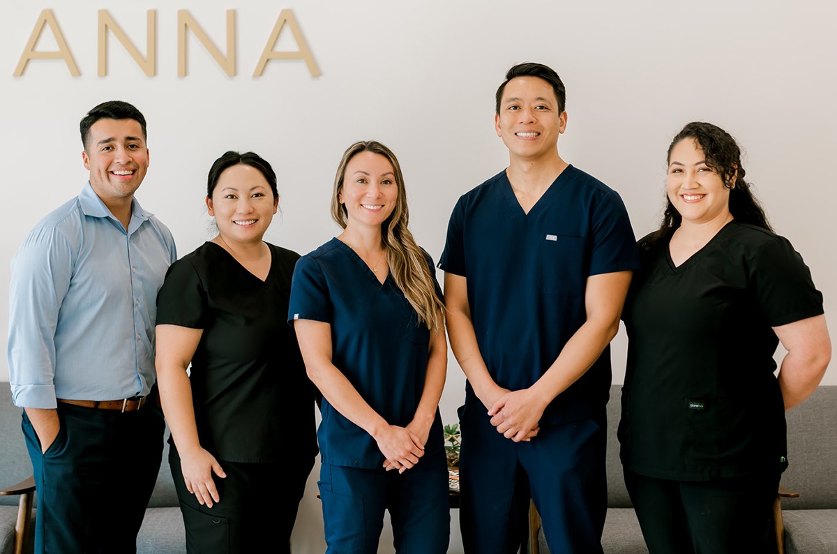 Five dental professionals pose together in front of ANNA branded wall in clinical setting.