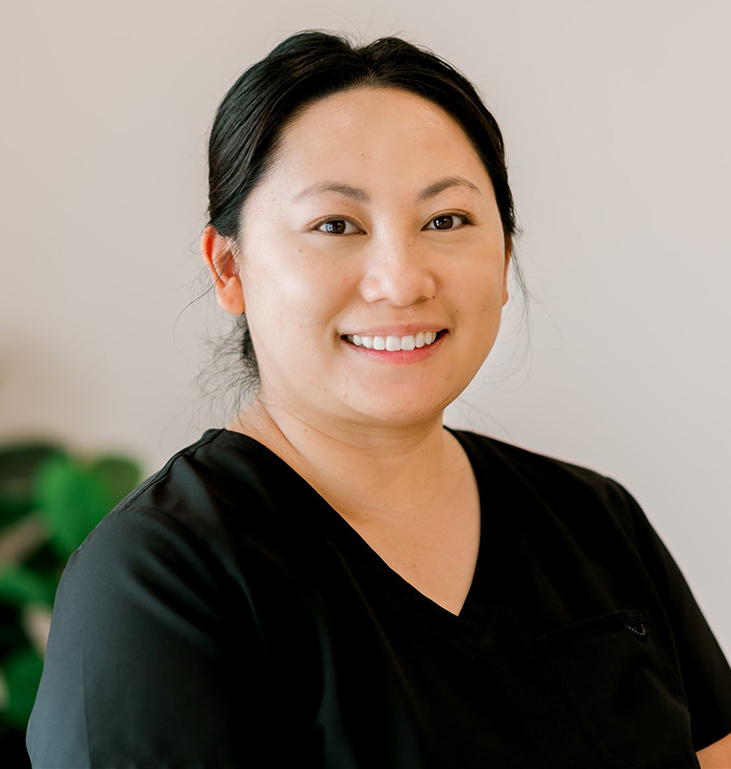 Smiling woman in black v-neck shirt against neutral background with green plant visible.