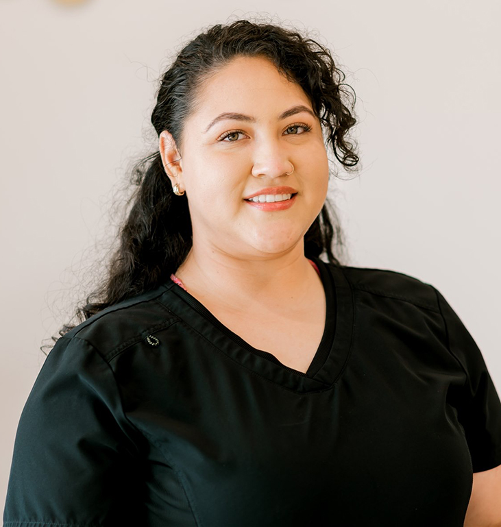 Professional headshot of woman smiling at camera against neutral background, wearing black scrubs.