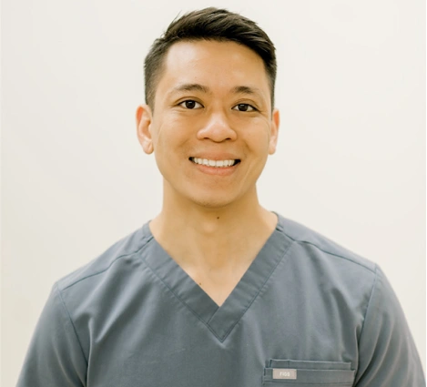Smiling male dental professional in gray scrubs against white background, headshot portrait.