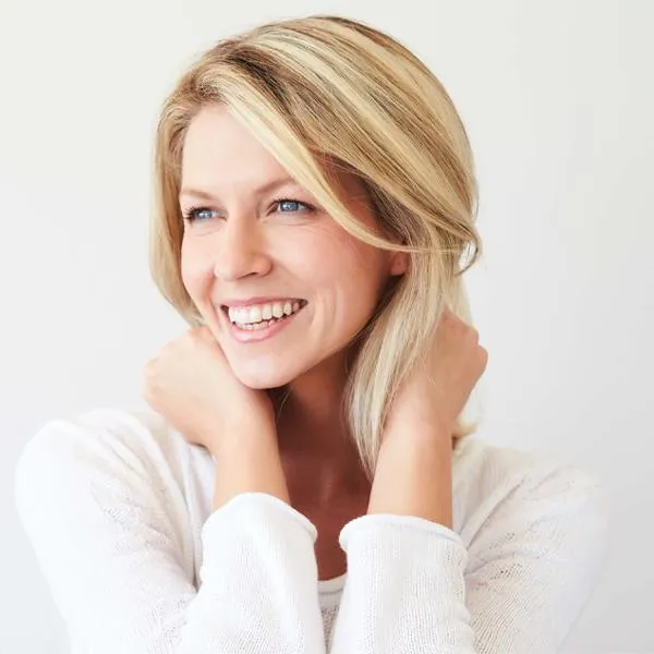 Smiling blonde woman in white sweater poses with hands near face against neutral background.